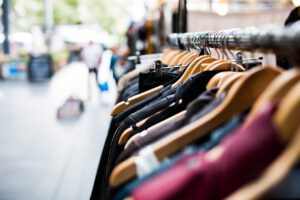 Kleiderstange mit verschiedenen Kleidungsstücken auf Holzkleiderbügeln in einem Straßenmarkt oder Outdoor-Shop, unscharfer Hintergrund mit Passanten.