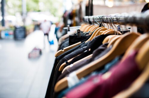 Kleiderstange mit verschiedenen Kleidungsstücken auf Holzkleiderbügeln in einem Straßenmarkt oder Outdoor-Shop, unscharfer Hintergrund mit Passanten.