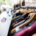 Kleiderstange mit verschiedenen Kleidungsstücken auf Holzkleiderbügeln in einem Straßenmarkt oder Outdoor-Shop, unscharfer Hintergrund mit Passanten.